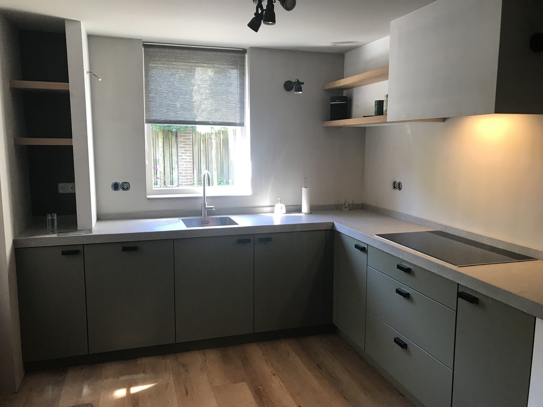 U-keuken met matte salie-grijze fronten, zwarte grepen, een werkblad in steenlook, inductiekookplaat en eiken wandplanken